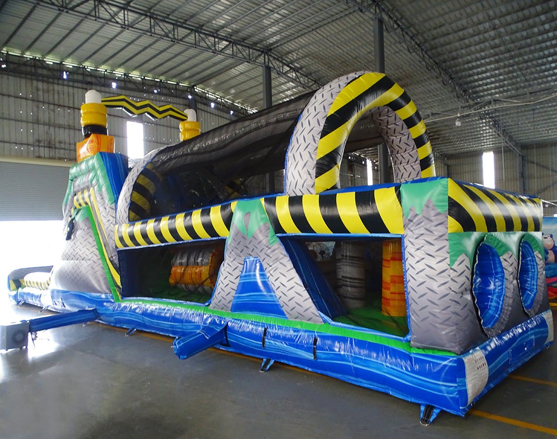 Wide-angle view of 53ft XL Radioactive Run Commercial Hybrid Inflatable Obstacle Course set up in a large indoor warehouse. The inflatable features yellow and black hazard stripes, multiple arches, tunnels, and obstacles. Metal roofing, support beams, and various other inflatable products are visible in the background, showcasing its size and commercial setting.