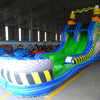 Front view of 53ft XL Radioactive Run Commercial Hybrid Inflatable Obstacle Course showing the entrance area. Features a large yellow and black striped arch, dual circular entrances with blue trim, and a diamond plate pattern base. The inflatable includes green slides and multiple obstacles. Set in a warehouse with concrete flooring and other inflatable products visible.