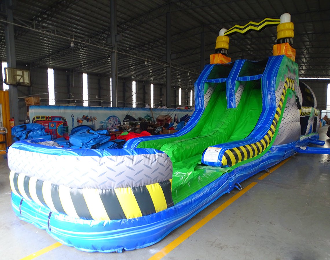 Front view of 53ft XL Radioactive Run Commercial Hybrid Inflatable Obstacle Course showing the entrance area. Features a large yellow and black striped arch, dual circular entrances with blue trim, and a diamond plate pattern base. The inflatable includes green slides and multiple obstacles. Set in a warehouse with concrete flooring and other inflatable products visible.