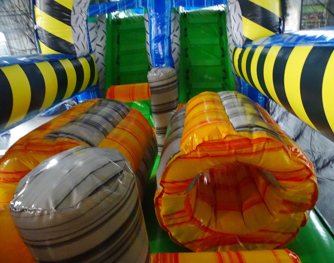 Interior view of 53ft XL Radioactive Run Commercial Hybrid Inflatable Obstacle Course showing inflatable obstacles. The image displays yellow and gray cylindrical barriers, green flooring, and yellow and black striped walls. Various inflatable obstacles are visible, demonstrating the course's challenging layout.