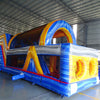 Three-quarter view of 53ft XL Rip Curl Commercial Hybrid Inflatable Obstacle Course displaying its full length. The structure has a blue and yellow design with dual red slides, multiple obstacles, and a large circular splash pool. Set in a large indoor warehouse with metal roofing and other inflatable products visible.
