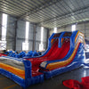 Side view of 53ft XL Rip Curl Commercial Hybrid Inflatable Obstacle Course featuring dual red slides emerging from a blue and yellow structure. The inflatable includes multiple obstacles, tunnels, and a large circular splash pool. Set up in a spacious warehouse with metal roofing and bright lighting visible.
