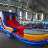 Side view of 53ft XL Rip Curl Commercial Hybrid Inflatable Obstacle Course featuring a blue and yellow color scheme. The inflatable structure includes dual red slides, multiple obstacles, and a large circular splash pool. Set up in a spacious warehouse with metal roofing and bright lighting visible.
