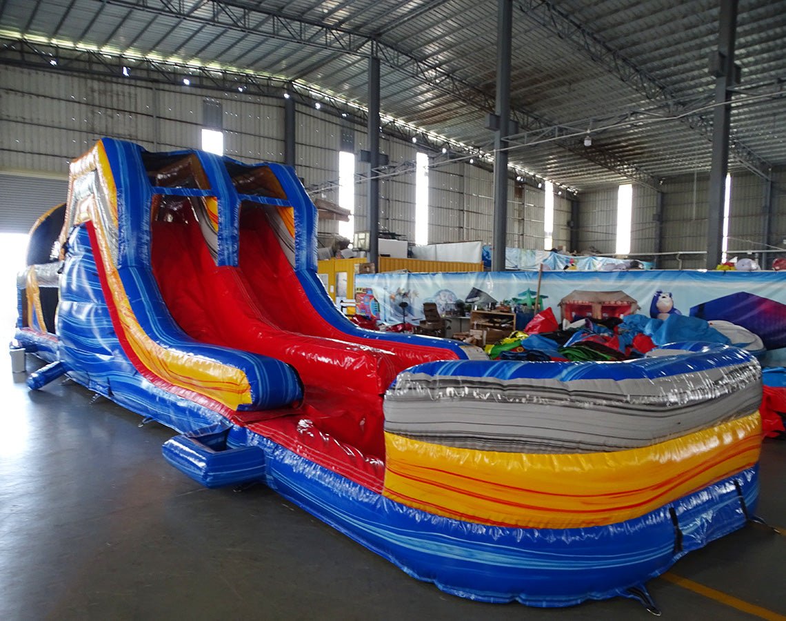 Side view of 53ft XL Rip Curl Commercial Hybrid Inflatable Obstacle Course featuring a blue and yellow color scheme. The inflatable structure includes dual red slides, multiple obstacles, and a large circular splash pool. Set up in a spacious warehouse with metal roofing and bright lighting visible.
