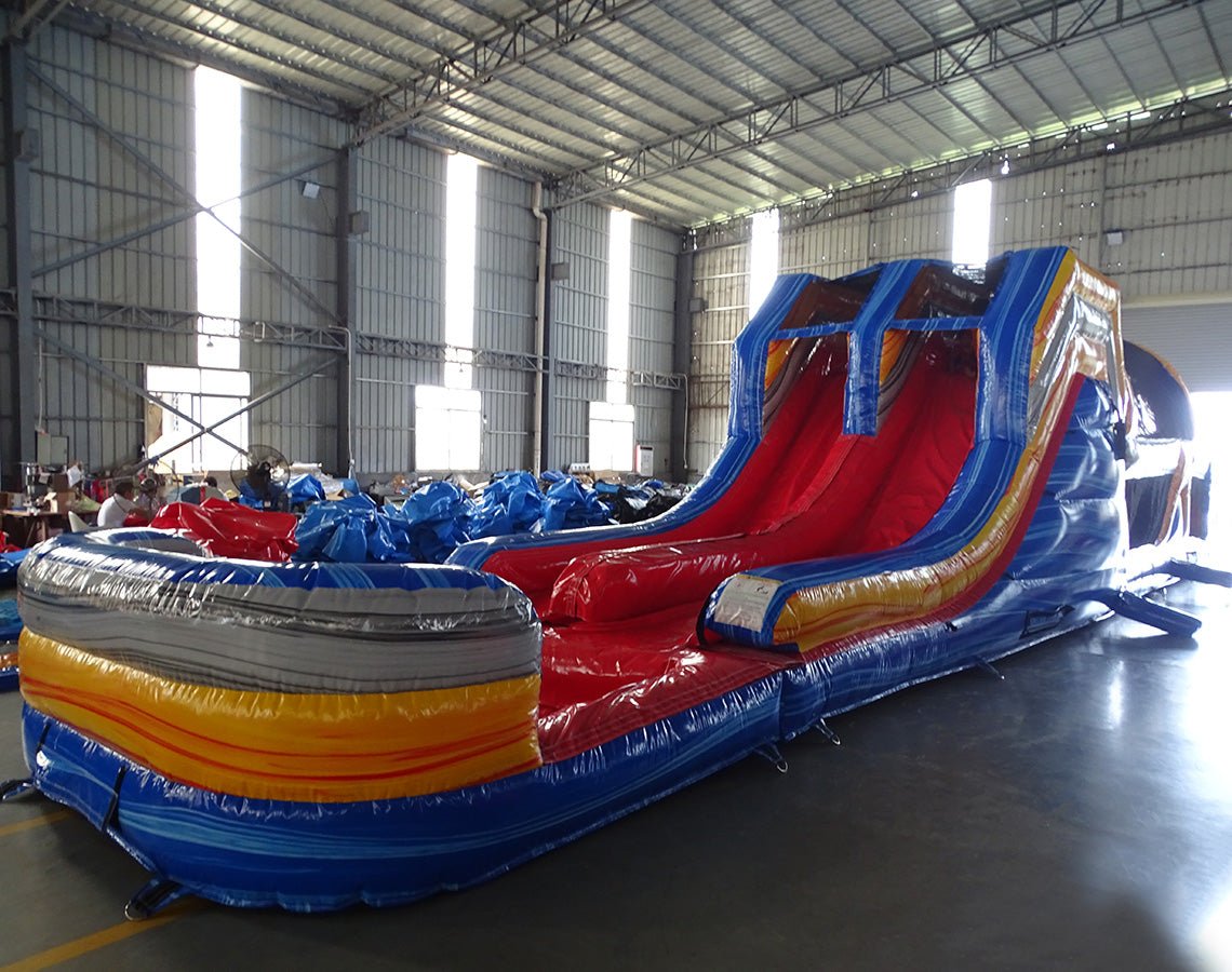 Front view of 53ft XL Rip Curl Commercial Hybrid Inflatable Obstacle Course showing dual red slides and a blue and yellow base. The inflatable features multiple obstacles, tunnels, and a large circular splash pool. Various other inflatable products and equipment are visible in the warehouse background.
