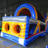 Back view of 53ft XL Rip Curl Commercial Hybrid Inflatable Obstacle Course showing dual circular entrances with a blue and yellow color scheme. The inflatable features a large arched top and multiple obstacles. Set in a warehouse with concrete flooring and various other inflatable products visible in the background.
