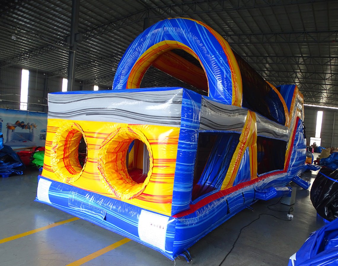 Back view of 53ft XL Rip Curl Commercial Hybrid Inflatable Obstacle Course showing dual circular entrances with a blue and yellow color scheme. The inflatable features a large arched top and multiple obstacles. Set in a warehouse with concrete flooring and various other inflatable products visible in the background.
