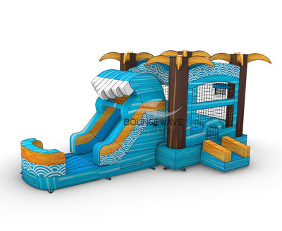 Wide-angle view of Akua Falls Commercial Econo Inflatable Combo (Wet/Dry) in a spacious warehouse. Shows the full structure including the blue wave-shaped body with white patterns, brown palm tree pillars, yellow fronds, curved slide, and splash pool. Warehouse interior with metal roofing, shelving units, and other inflatable products visible, providing scale context.