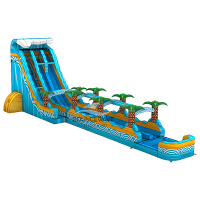 Large inflatable water slide with tropical theme, featuring bright blue color, palm tree decorations, and a long slip and slide extension. The main slide has two lanes and a tall climbing wall. Set up in an indoor warehouse with metal roofing and other inflatable products visible in the background.