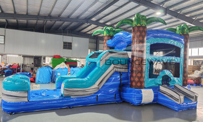 Tropical-themed inflatable combo with bounce house, slide, and splash pool. Decorated with palm trees and featuring blue and turquoise colors. Displayed in an indoor facility with various other inflatable products and equipment visible.