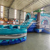 Wide-angle view of Bahama Curved Dual Lane Commercial Inflatable Combo (Wet/Dry) in a spacious warehouse. Features the full structure including the wave-patterned climbing wall, dual blue curved slides, inflatable palm trees, and large circular splash pool. Warehouse interior visible with metal roofing, shelving units, and other inflatable products, providing scale and context for the combo's size.