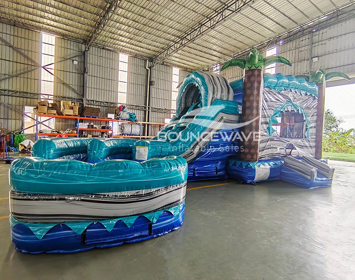 Wide-angle view of Bahama Curved Dual Lane Commercial Inflatable Combo (Wet/Dry) in a spacious warehouse. Features the full structure including the wave-patterned climbing wall, dual blue curved slides, inflatable palm trees, and large circular splash pool. Warehouse interior visible with metal roofing, shelving units, and other inflatable products, providing scale and context for the combo's size.