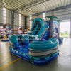 Side view of Bahama Curved Dual Lane Commercial Inflatable Combo (Wet/Dry) set up in a large indoor warehouse. Features dual blue curved slides, inflatable palm trees, and a tall climbing structure with wave patterns. The large circular splash pool area is prominent in the foreground. Metal roofing, shelving units, and other warehouse equipment visible, demonstrating its size in a commercial setting.