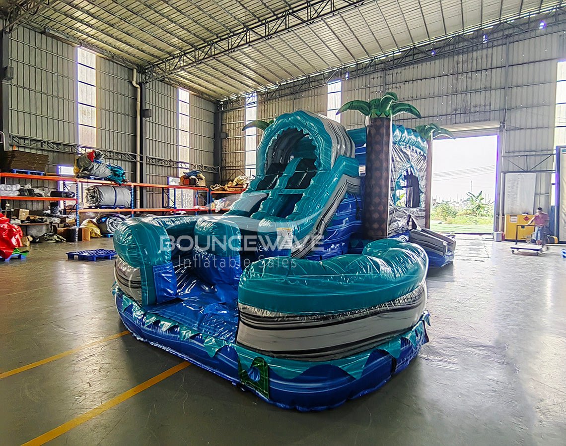 Side view of Bahama Curved Dual Lane Commercial Inflatable Combo (Wet/Dry) set up in a large indoor warehouse. Features dual blue curved slides, inflatable palm trees, and a tall climbing structure with wave patterns. The large circular splash pool area is prominent in the foreground. Metal roofing, shelving units, and other warehouse equipment visible, demonstrating its size in a commercial setting.