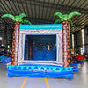 Large tropical-themed inflatable combo featuring a tall wave-patterned climbing wall leading to dual curved blue water slides. The structure includes inflatable palm trees on both sides, a circular splash pool area with wave designs, and a bounce house section. Set up in a spacious warehouse with metal roofing and shelving units visible in the background. The "BOUNCEWAVE Inflatable Sales" logo is prominently displayed on the inflatable.