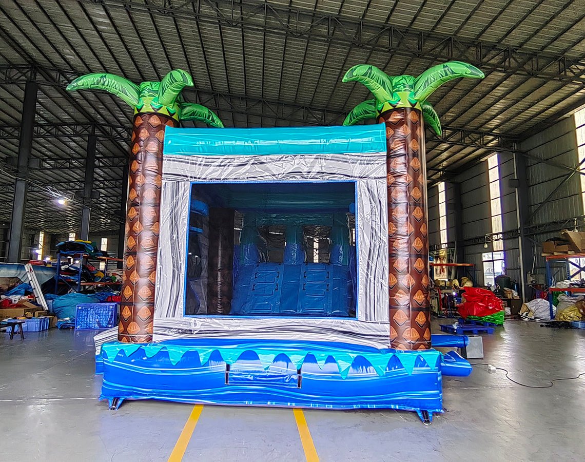 Large tropical-themed inflatable combo featuring a tall wave-patterned climbing wall leading to dual curved blue water slides. The structure includes inflatable palm trees on both sides, a circular splash pool area with wave designs, and a bounce house section. Set up in a spacious warehouse with metal roofing and shelving units visible in the background. The "BOUNCEWAVE Inflatable Sales" logo is prominently displayed on the inflatable.