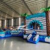 Side view of Bahama Curved Dual Lane Commercial Inflatable Combo (Wet/Dry) in an indoor warehouse. Highlights the wave-patterned climbing wall, dual blue curved slides, inflatable palm trees, and circular splash pool. Other inflatable products, storage containers, and warehouse equipment visible in the background, showcasing its scale in a commercial environment.