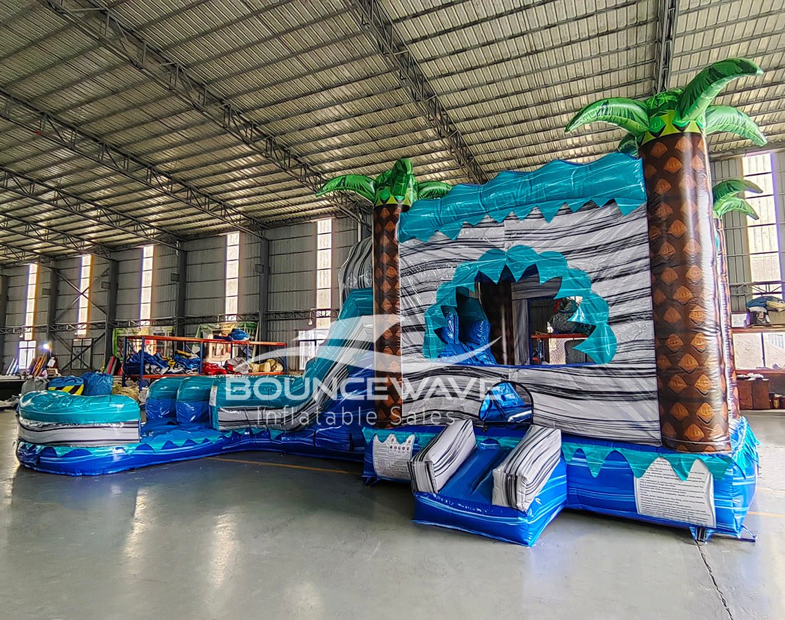 Side view of Bahama Curved Dual Lane Commercial Inflatable Combo (Wet/Dry) in an indoor warehouse. Highlights the wave-patterned climbing wall, dual blue curved slides, inflatable palm trees, and circular splash pool. Other inflatable products, storage containers, and warehouse equipment visible in the background, showcasing its scale in a commercial environment.