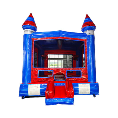 Inflatable bounce house with castle theme, featuring blue walls, red turrets, and a red entrance ramp. The structure has two spired towers and a large opening with mesh windows. Safety instructions are visible on the front panels. Set up indoors with a window visible in the background.