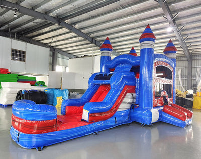 Large inflatable castle combo with bounce house, slide, and splash pool. Features blue and red colors with castle-like design elements. Set up in an indoor warehouse with metal roofing and other inflatable products visible in the background.