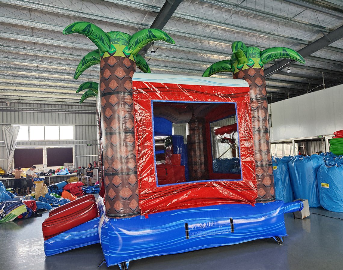 Inflatable tropical-themed combo featuring a bounce house with palm tree decorations, attached slide, and splash pool. The structure has vibrant red, blue, and white colors with inflatable palm trees. Set up in an indoor warehouse with metal roofing and other equipment visible in the background.