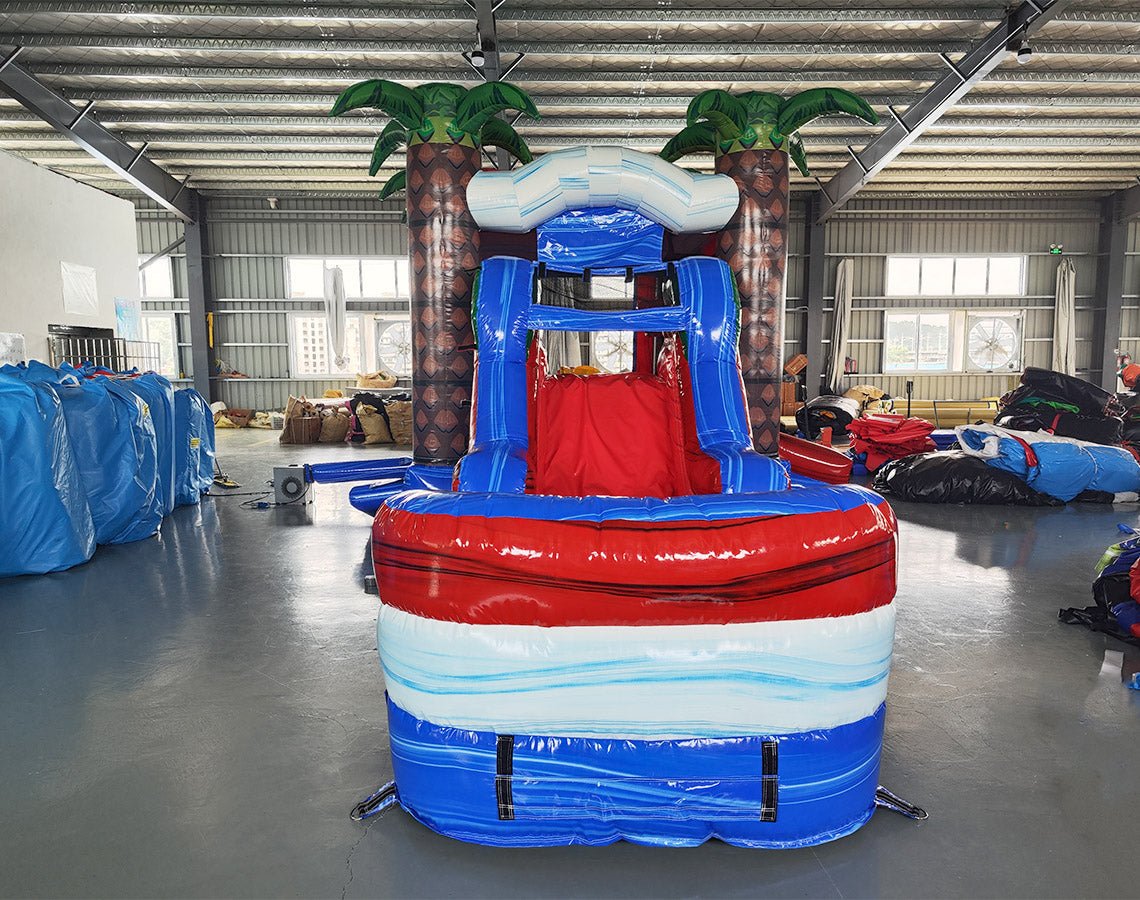 Front view of Baja Thunder Econo Combo, showcasing the bounce house entrance flanked by inflatable palm trees, with a red slide leading to a blue and white splash pool. Set up in an indoor facility with other inflatable products visible in the background.