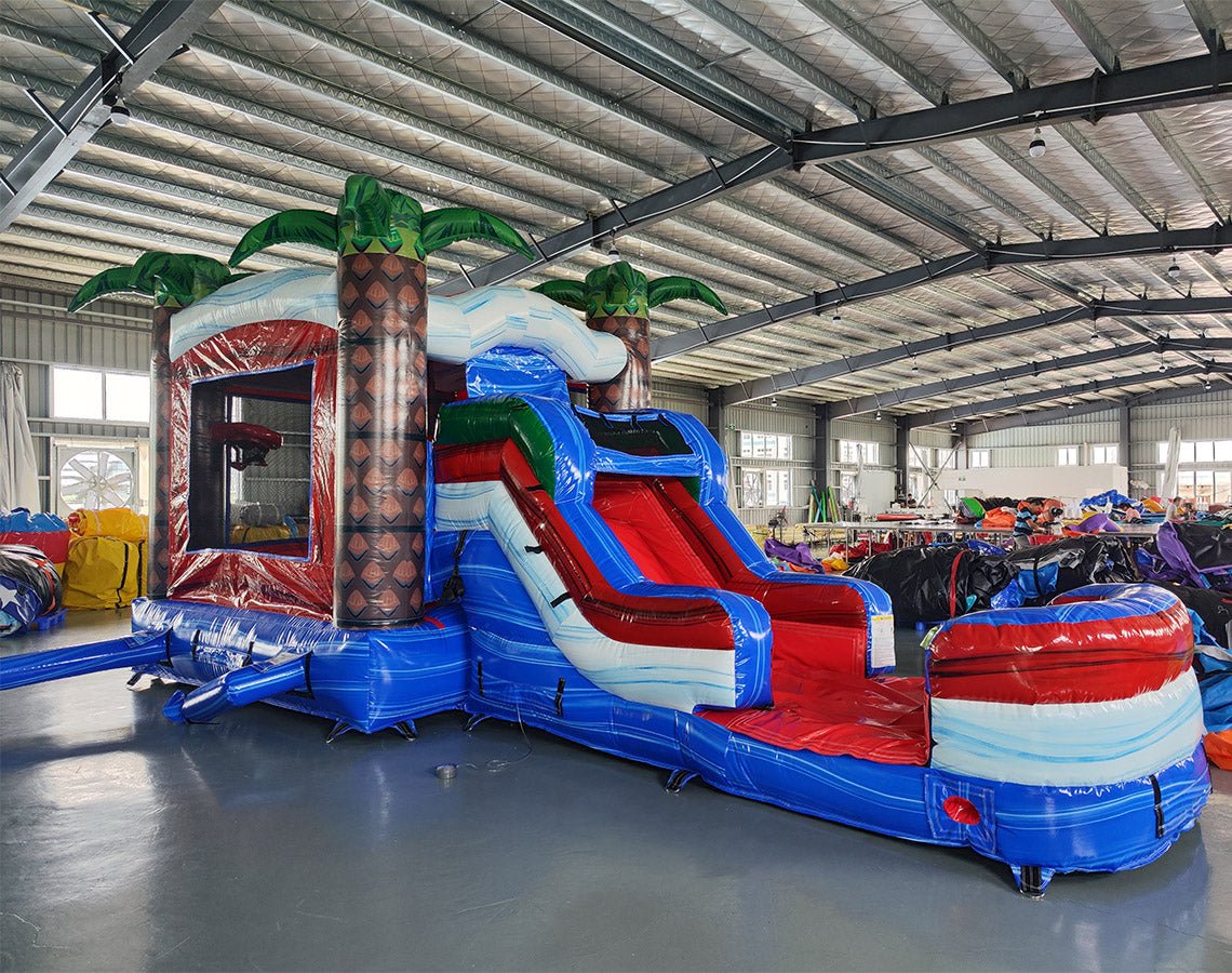 Inflatable tropical-themed combo featuring a bounce house with palm tree decorations, attached slide, and splash pool. The structure has vibrant blue, red, and white colors with brown palm tree trunks. Set up in an indoor warehouse with metal roofing visible.