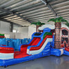 Angled view of the Baja Thunder Econo Combo featuring a blue and white bounce house with palm tree decorations, red slide, and spacious splash pool. The inflatable is set up in an indoor warehouse with other equipment and inflatables visible.