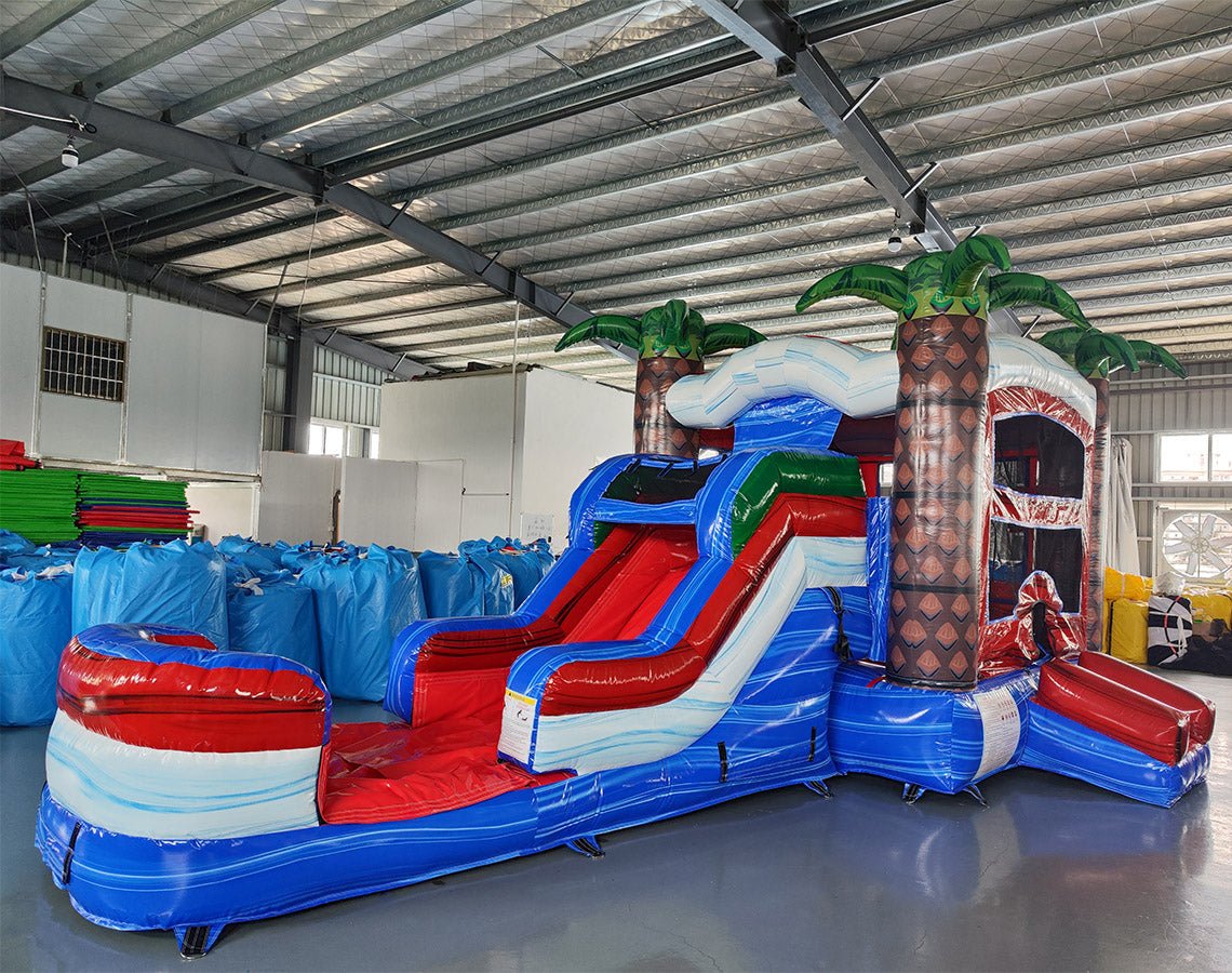 Angled view of the Baja Thunder Econo Combo featuring a blue and white bounce house with palm tree decorations, red slide, and spacious splash pool. The inflatable is set up in an indoor warehouse with other equipment and inflatables visible.