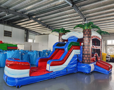 Angled view of the Baja Thunder Econo Combo featuring a blue and white bounce house with palm tree decorations, red slide, and spacious splash pool. The inflatable is set up in an indoor warehouse with other equipment and inflatables visible.