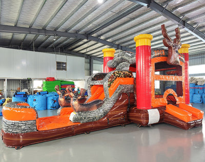 Angled view of the Buck Shot Econo Combo featuring an orange and brown bounce house with deer decorations, red pillars, and tree trunk obstacles. The inflatable is set up in an indoor warehouse with other inflatable products and equipment visible around it.