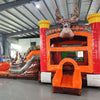 Side view of Buck Shot Econo Combo showing bright orange, red, and brown design with inflatable deer figures and tree trunk obstacles. The combo includes a bounce house, slide, and climbing elements. Displayed in an indoor facility with other inflatables visible.