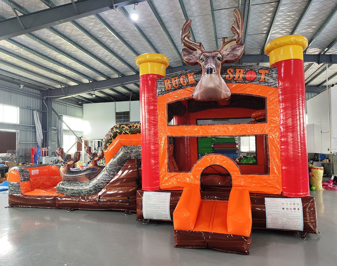 Side view of Buck Shot Econo Combo showing bright orange, red, and brown design with inflatable deer figures and tree trunk obstacles. The combo includes a bounce house, slide, and climbing elements. Displayed in an indoor facility with other inflatables visible.