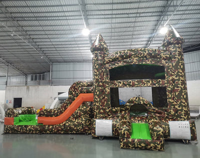 Angled view of the Camo Econo Combo featuring a camouflage-patterned bounce house with turrets, orange slide, and green splash pool. The inflatable is set up in an indoor warehouse with metal roofing and other equipment visible in the background.