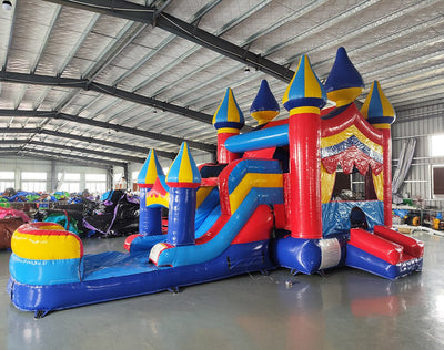 Wide-angle view of the Carnival Econo Combo featuring a colorful bounce house with multiple spires, attached slide, and large splash pool area. The inflatable is set up in a spacious indoor warehouse with metal roofing, bright lighting, and other inflatable products visible in the background.