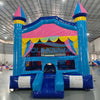 Front view of Carnival Pink Commercial Grade Bounce House set up in a large indoor warehouse. Features a vibrant blue castle-shaped structure with pink and yellow accents. Three turrets with blue and pink conical tops adorn the bounce house. The entrance has a yellow scalloped awning and mesh windows. A blue slide is attached to the side. The "BOUNCEWAVE" logo is visible on the front.