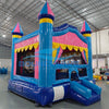 Side view of Carnival Pink Commercial Grade Bounce House in a warehouse setting. Highlights the blue castle structure with pink walls and yellow trim. Shows the full length of the attached blue slide and the three turrets with blue and pink conical tops. Mesh windows and a yellow scalloped awning are visible. Other inflatable products and warehouse equipment can be seen in the background.