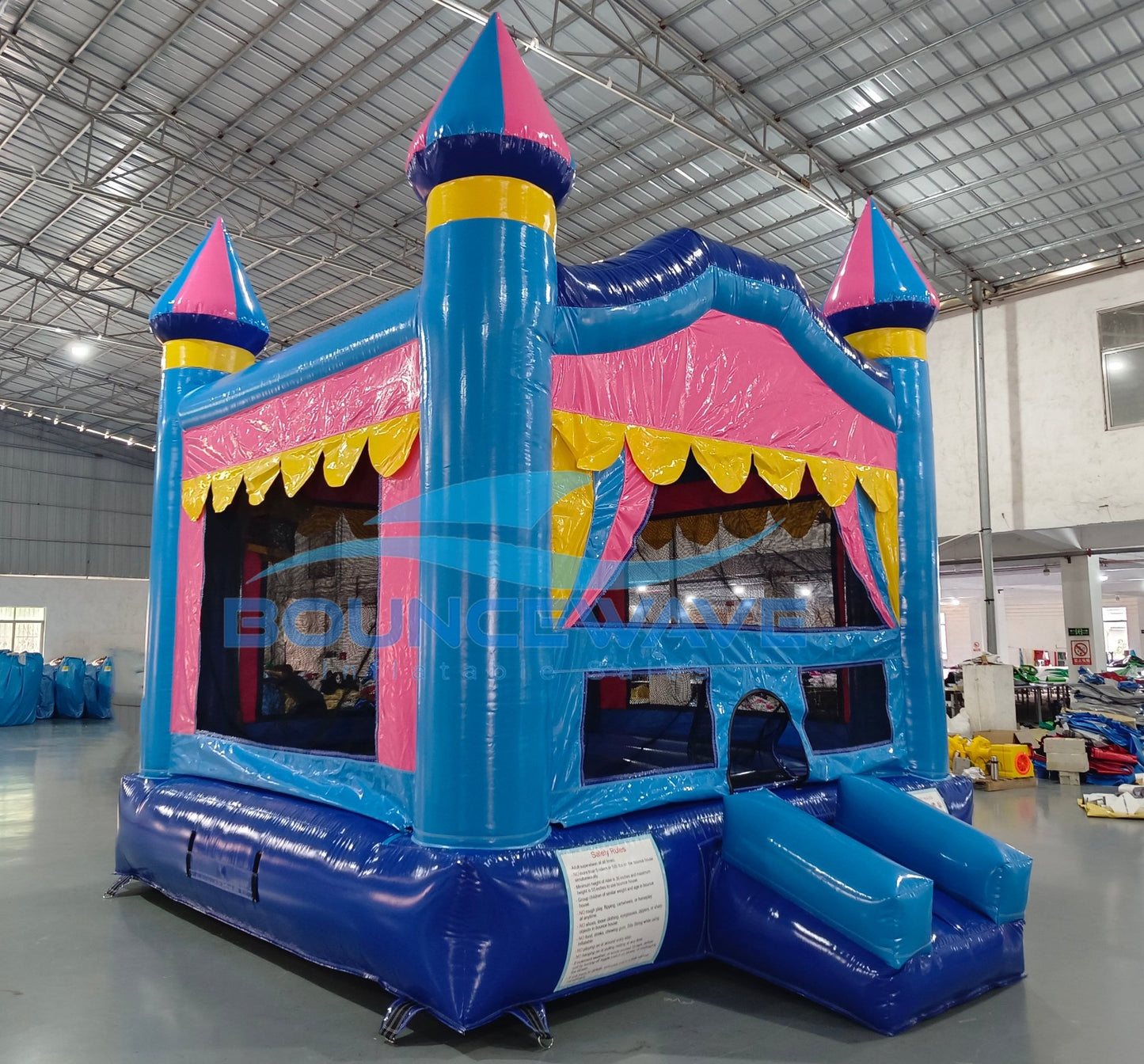 Side view of Carnival Pink Commercial Grade Bounce House in a warehouse setting. Highlights the blue castle structure with pink walls and yellow trim. Shows the full length of the attached blue slide and the three turrets with blue and pink conical tops. Mesh windows and a yellow scalloped awning are visible. Other inflatable products and warehouse equipment can be seen in the background.