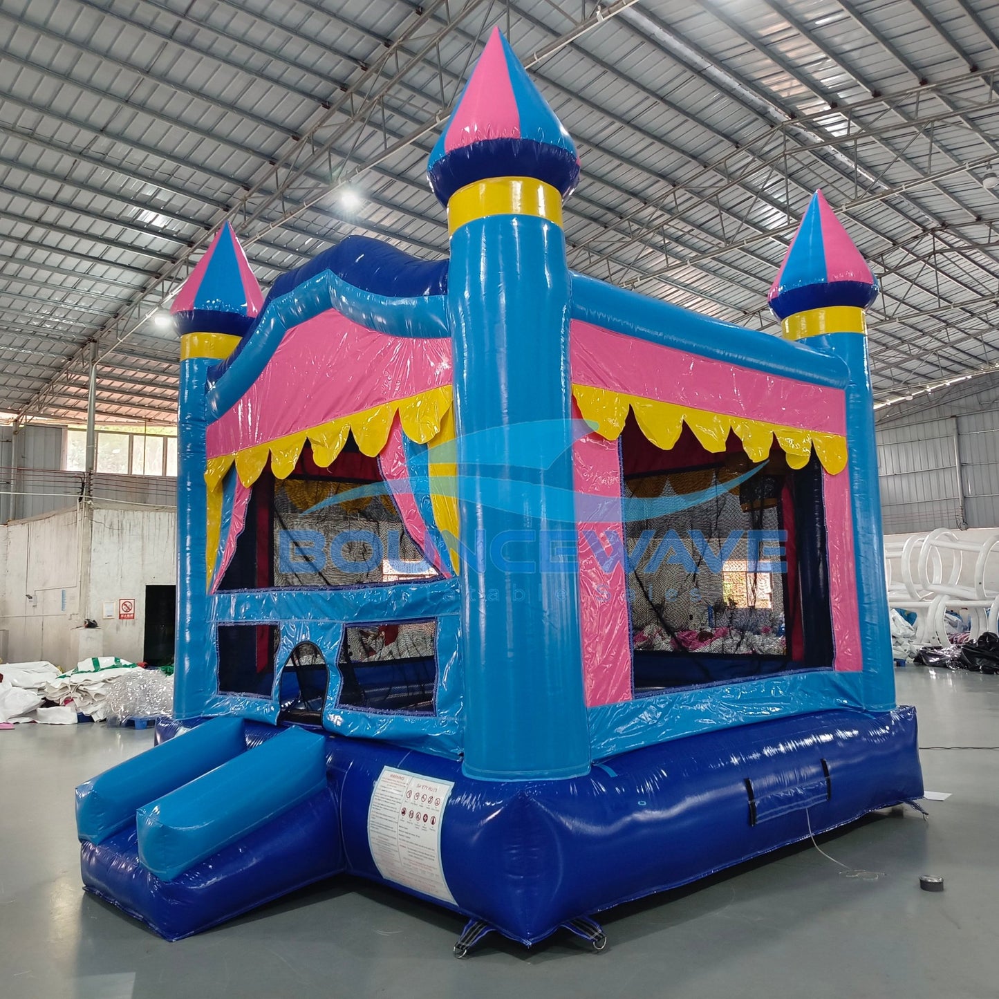 Three-quarter view of Carnival Pink Commercial Grade Bounce House in a warehouse. Showcases the blue castle structure with pink walls, yellow trim, and three turrets with blue and pink conical tops. The blue slide, mesh windows, and yellow scalloped awning over the entrance are visible. The image provides a good sense of the bounce house's size relative to the warehouse space.