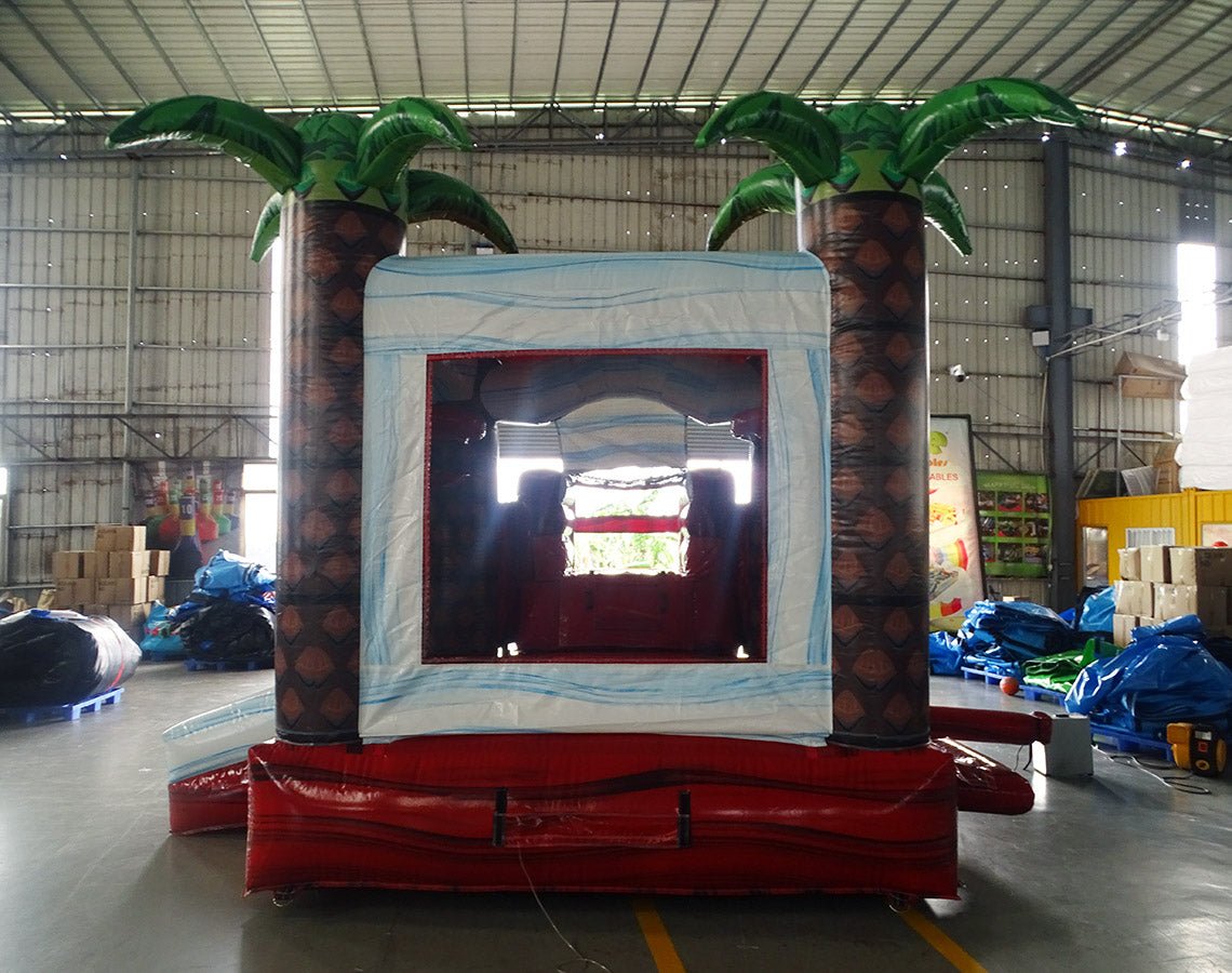 Large inflatable tropical-themed combo featuring a bounce house with palm tree decorations, attached slide, and splash pool. The structure has vibrant red and white colors with brown palm tree trunks. Set up in an indoor warehouse with metal roofing and other inflatable products visible in the background.