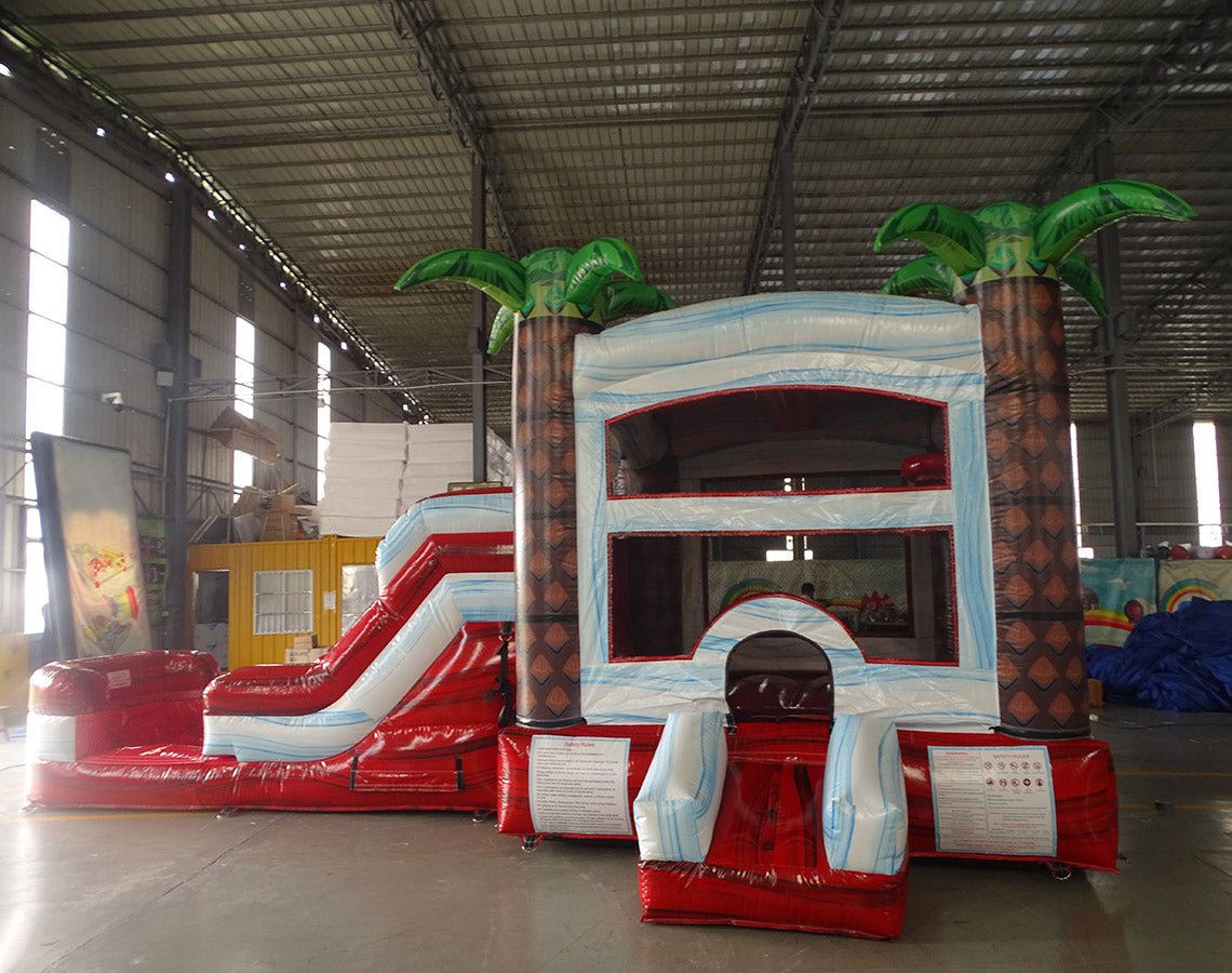 Front view of Crimson Bay Econo Combo, showcasing the bounce house entrance with palm tree decorations, red slide, and white splash pool area. Set up in an indoor facility with other inflatable products and equipment visible in the background.