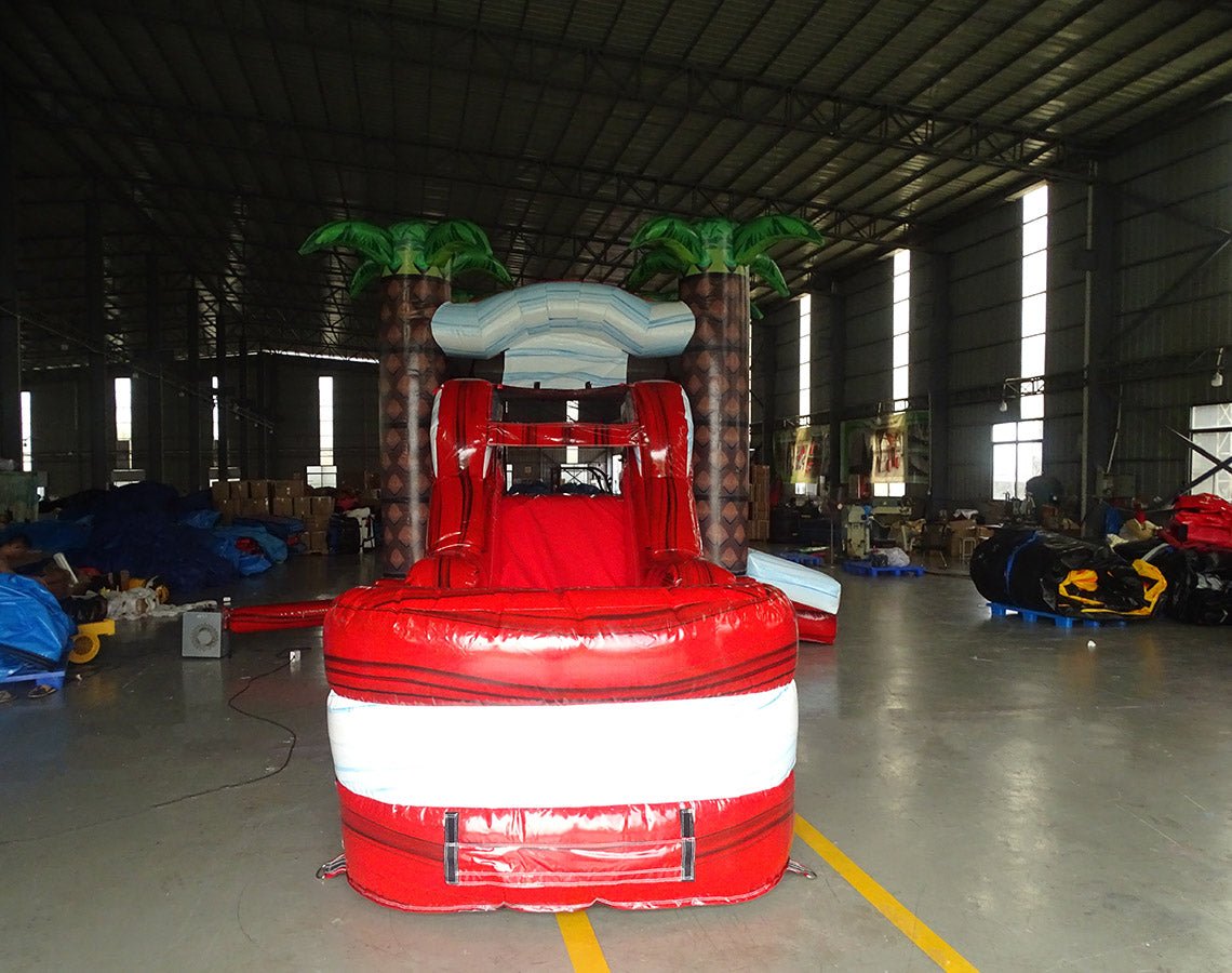 Large inflatable tropical-themed combo featuring a bounce house with palm tree decorations, attached slide, and splash pool. The structure has vibrant red and white colors with brown palm tree trunks. Set up in an indoor warehouse with metal roofing visible.