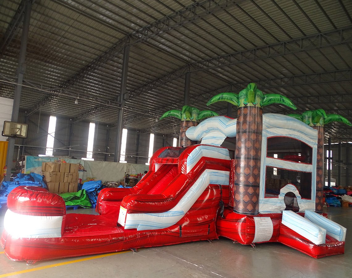 Side angle view of the Crimson Bay Econo Combo featuring a red and white bounce house with palm tree decorations, curved slide, and spacious splash pool. The inflatable is set up in an indoor warehouse with other equipment and inflatables visible.