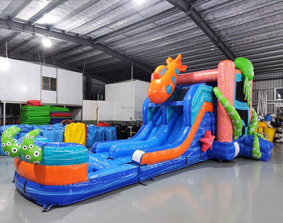 Wide-angle view of Deep Sea Dive Dual Lane Commercial Inflatable Combo in a spacious warehouse. The inflatable features dual blue slides, a large orange octopus, green tentacles, and a blue wave-patterned base with a splash pool. Metal roofing, support beams, and other inflatable products visible in the background, demonstrating its size and commercial setting.