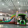 Wide-angle view of Dino Dive Dual Lane Commercial Inflatable Combo in a large indoor warehouse. Features a gray castle structure with green accents, flame-topped towers, and two large T-Rex figures flanking the entrance. Dual slides, inflatable vegetation, and a splash pool area are visible. Metal roofing, support beams, and other inflatable products in the background showcase the commercial setting.