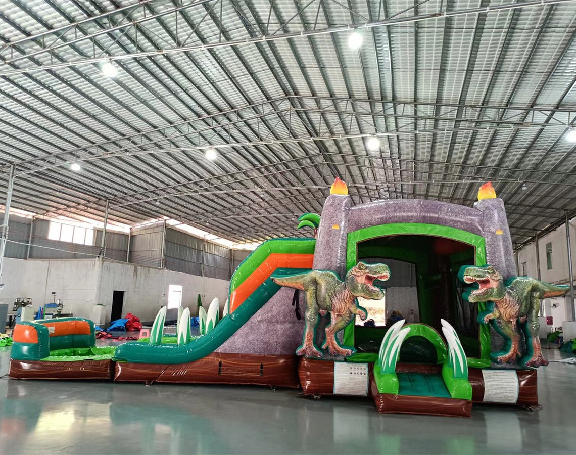 Wide-angle view of Dino Dive Dual Lane Commercial Inflatable Combo in a large indoor warehouse. Features a gray castle structure with green accents, flame-topped towers, and two large T-Rex figures flanking the entrance. Dual slides, inflatable vegetation, and a splash pool area are visible. Metal roofing, support beams, and other inflatable products in the background showcase the commercial setting.