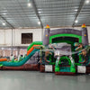 Wide-angle view of Dino Dive Dual Lane Commercial Inflatable Combo in a spacious warehouse. The inflatable features a gray castle structure with green accents, flame-topped towers, and two large T-Rex figures. Dual slides, bounce area, and inflatable vegetation are visible. Metal roofing, support beams, and other warehouse equipment provide scale context.