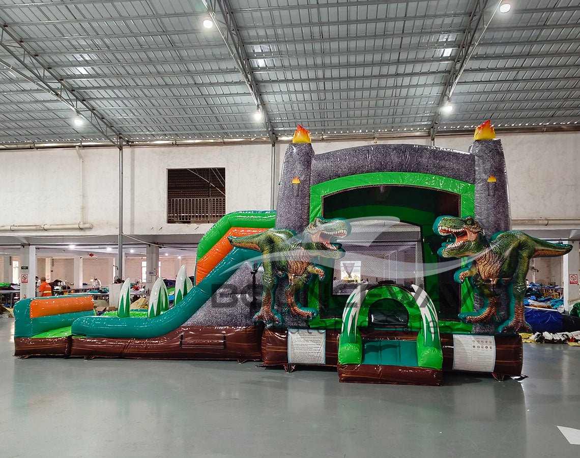 Wide-angle view of Dino Dive Dual Lane Commercial Inflatable Combo in a spacious warehouse. The inflatable features a gray castle structure with green accents, flame-topped towers, and two large T-Rex figures. Dual slides, bounce area, and inflatable vegetation are visible. Metal roofing, support beams, and other warehouse equipment provide scale context.