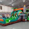 Front angled view of Dino Dive Dual Lane Commercial Inflatable Combo in a warehouse environment. Highlights the gray castle structure with green trim, flame-topped towers, and two imposing T-Rex figures. The dual slides, bounce area, and inflatable foliage are prominently displayed. Other inflatable products and warehouse equipment visible, showcasing its size and commercial application.