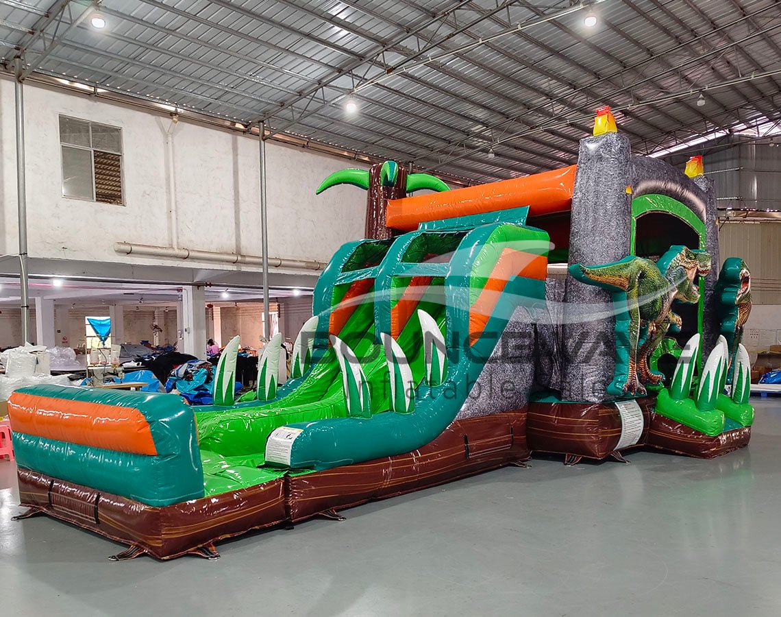 Front angled view of Dino Dive Dual Lane Commercial Inflatable Combo in a warehouse environment. Highlights the gray castle structure with green trim, flame-topped towers, and two imposing T-Rex figures. The dual slides, bounce area, and inflatable foliage are prominently displayed. Other inflatable products and warehouse equipment visible, showcasing its size and commercial application.